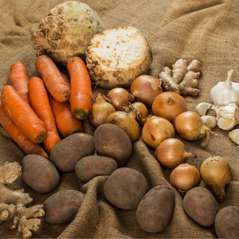 A selection of vegetables, including carrots, potatoes, onions, garlic, ginger, and celeriac, arranged on a burlap cloth.