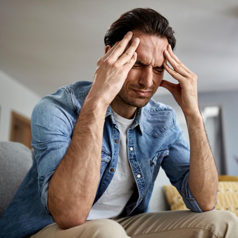 A man in a denim shirt sitting on a couch with his hands on his temples, appearing to be in pain or stressed.