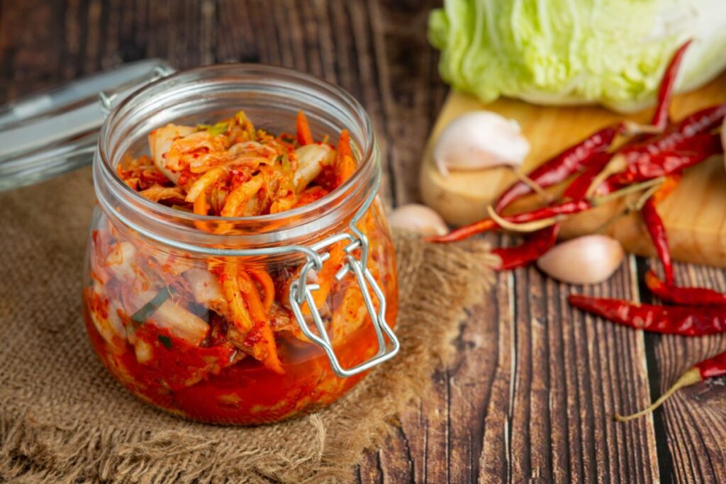A glass jar of kimchi is placed on a burlap cloth. In the background, there are garlic bulbs, red chili peppers, and a wedge of napa cabbage on a wooden surface.