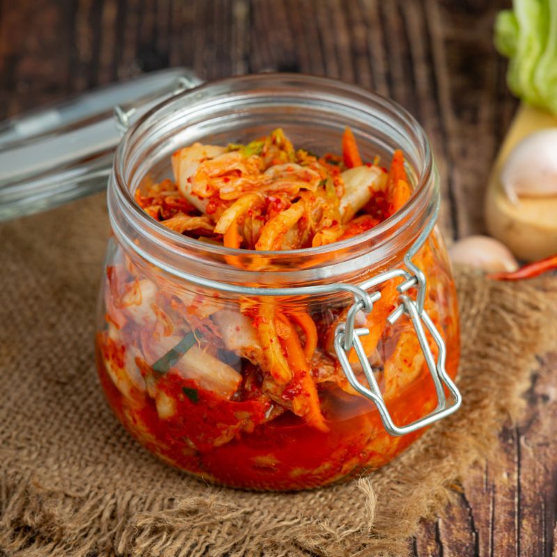 A jar of homemade kimchi with a metal latch on a burlap cloth, surrounded by fresh ingredients on a wooden surface.