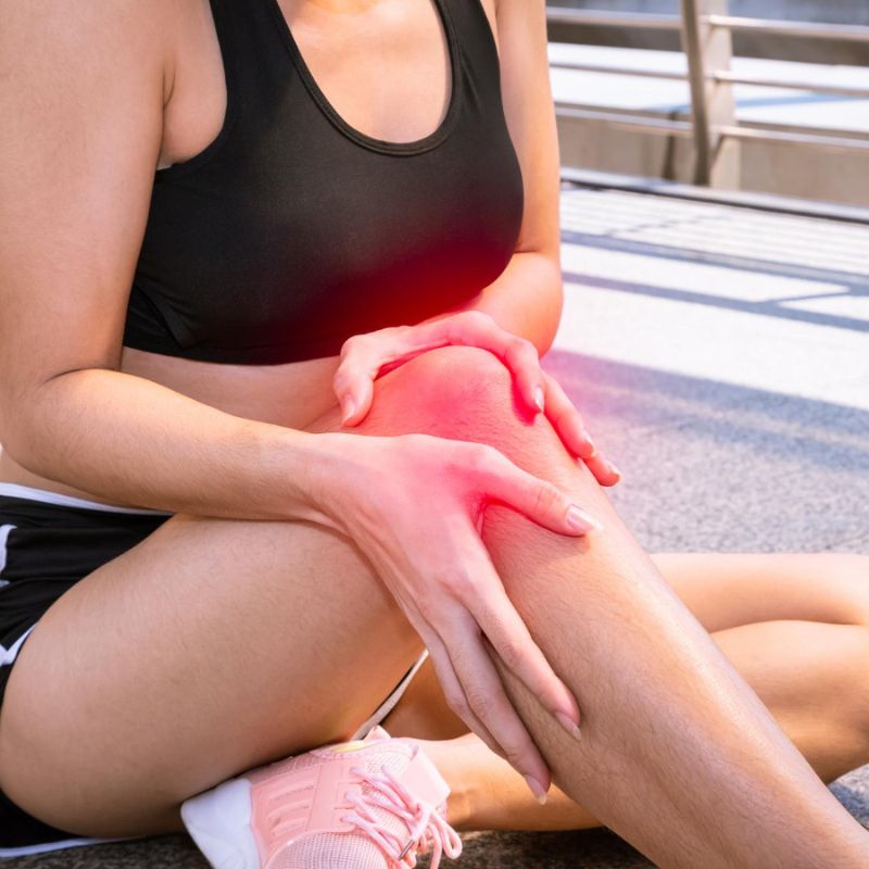 Person in athletic wear holding their injured knee, highlighted in red, while sitting on the ground. The knee looks inflamed.
