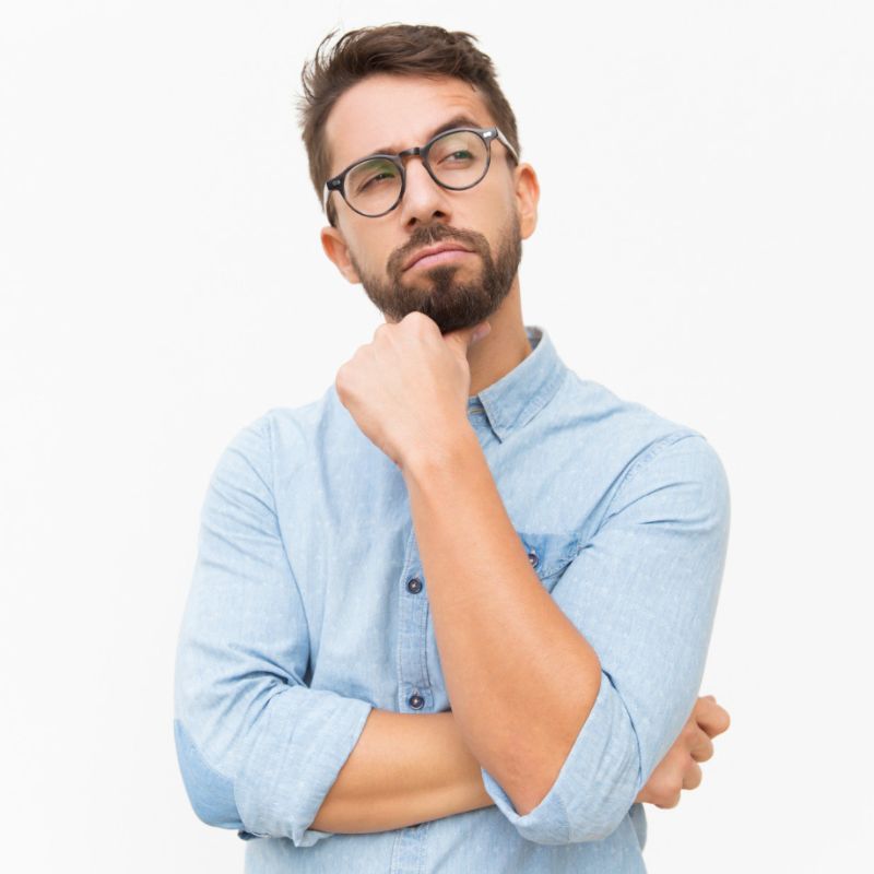 A man with glasses and a beard, wearing a light blue shirt, stands thoughtfully with his hand on his chin against a plain white background.