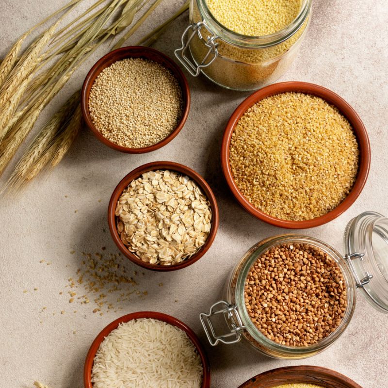 Assorted grains and cereals, including quinoa, oats, millet, and rice, are displayed in brown bowls and glass jars alongside wheat stalks on a light-colored surface—perfect for those following an anti candida diet.
