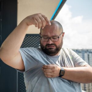 A man wearing glasses and a gray T-shirt is outdoors, looking at a large sweat stain under his arm with a concerned expression, wondering if it might be a symptom of nonalcoholic fatty liver disease.