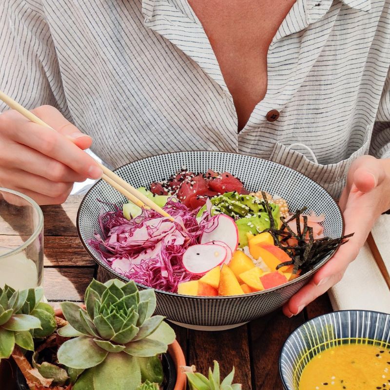 Person in a striped shirt holding chopsticks with a bowl of mixed salad, including radishes, cabbage, avocado, and seaweed, on a wooden table beside succulents and a small dish of yellow sauce—perfect for those seeking candida diet recipes.