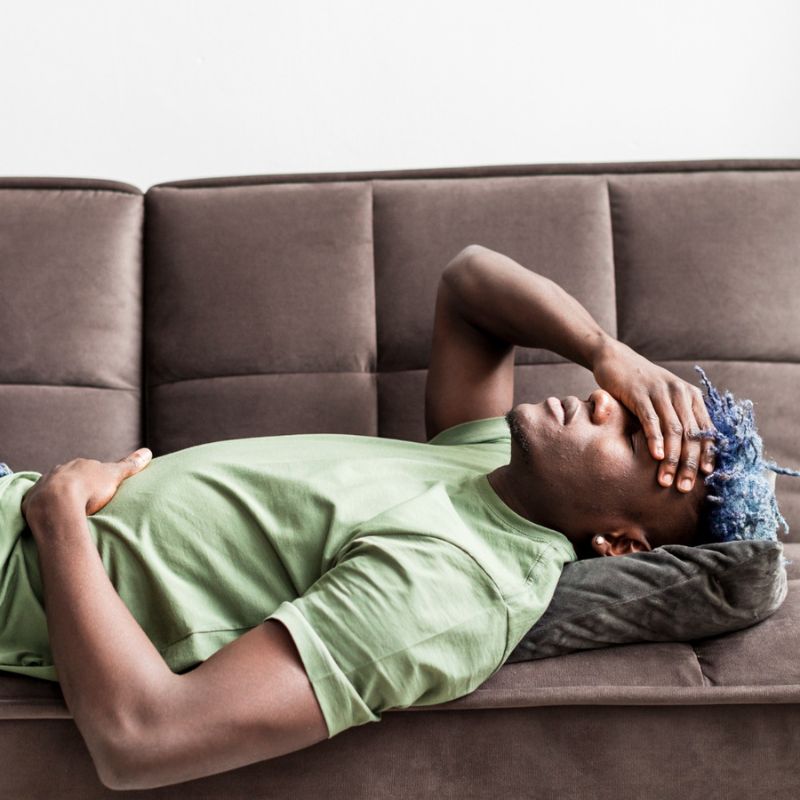 Person lying on a sofa with one hand on their forehead and the other on their stomach, wearing a green shirt and with blue hair. The person is suffering with the effects of Candida die off.