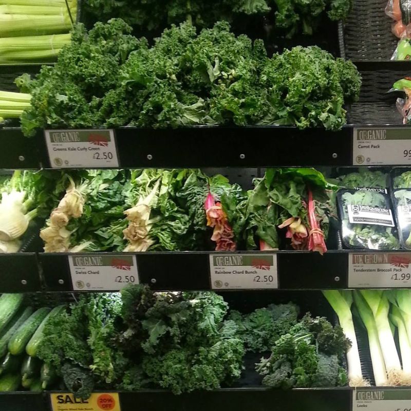 A supermarket shelf displays various leafy greens, including green kale, rainbow chard, Swiss chard, and tenderstem broccoli—ideal choices for those on an anti candida diet. Price tags and labels indicating organic options are visible.