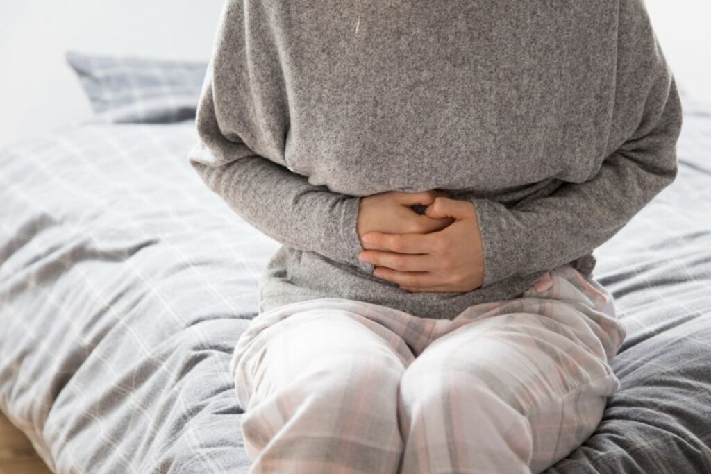 A person sits on a bed, wearing a grey sweater and checkered pajama pants, holding their stomach with both hands, suggesting abdominal discomfort—perhaps they’re considering starting a candida detox diet to find some relief.