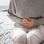 A person sits on a bed, wearing a grey sweater and checkered pajama pants, holding their stomach with both hands, suggesting abdominal discomfort—perhaps they’re considering starting a candida detox diet to find some relief.