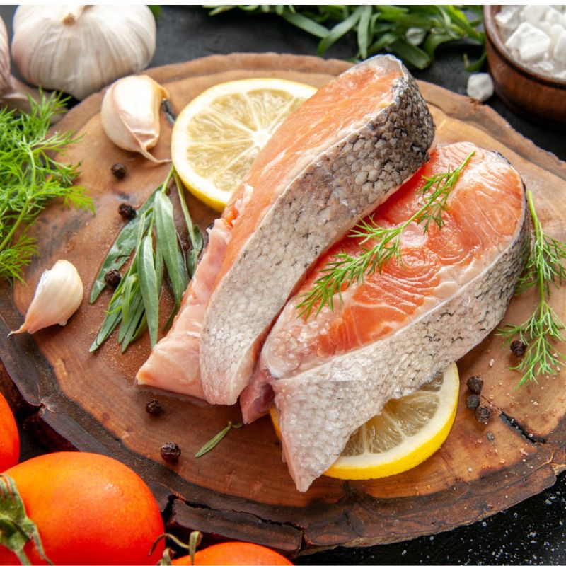         Two raw salmon fillets garnished with dill and tarragon sit on a wooden cutting board with lemon slices, surrounded by garlic cloves, cherry tomatoes, and peppercorns, showcasing the rich oils and fats that play essential functions in the body.