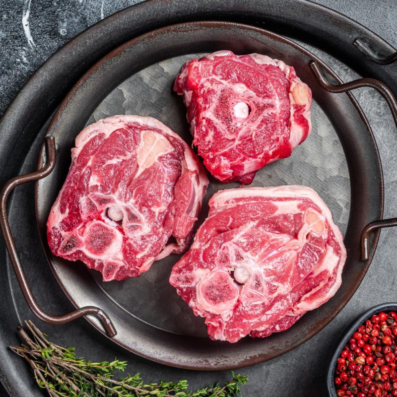 Three raw meat cuts arranged in a circular black dish, garnished with thyme sprigs and whole pink peppercorns on the side, a drizzle of olive oil enhancing their flavors and adding beneficial fats essential for the body's functions.