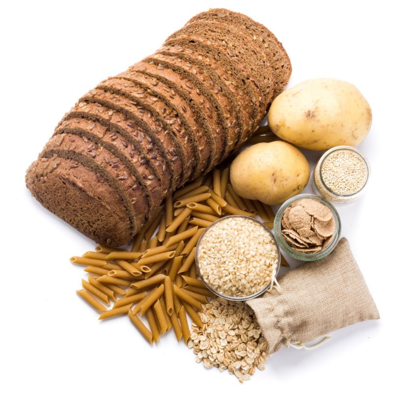 A variety of carbohydrate foods including sliced whole grain bread, potatoes, pasta, oats, quinoa, rice, and wheat bran.