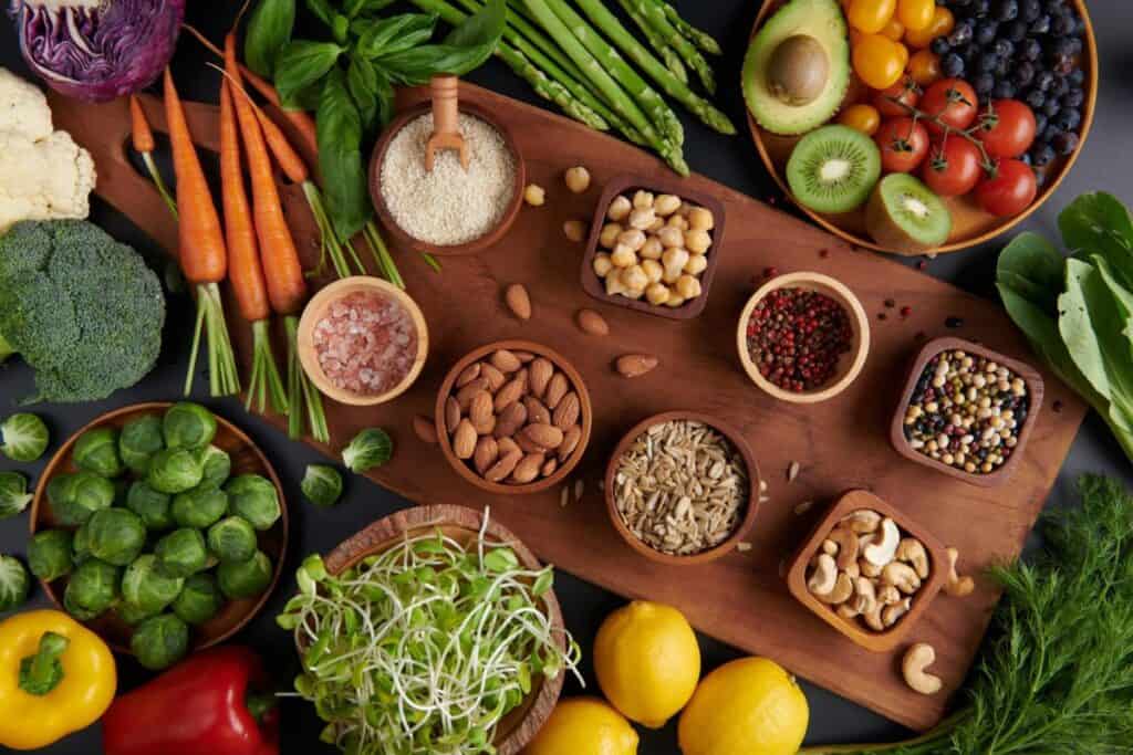 An assortment of vegetables, fruits, nuts, seeds, and spices on a wooden board. Items include carrots, cauliflower (rich in complex carbohydrates), tomatoes, kiwi, brussels sprouts, lemons, and various bowls of grains and nuts.