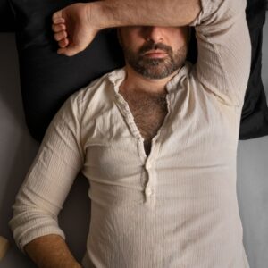 A bearded man in a light-colored shirt is lying down with one arm covering his forehead, seemingly exhausted from taking a candida saliva test.