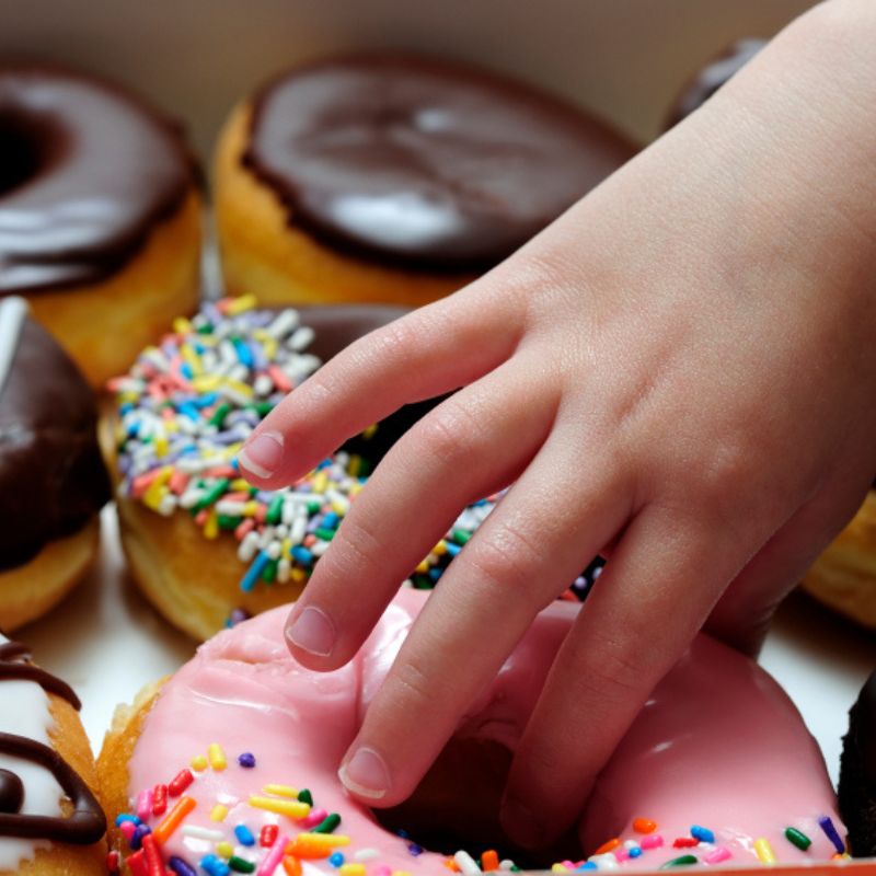 A child's hand reaches for a pink frosted donut with colorful sprinkles, among an assortment of chocolate-topped donuts—a sweet escape from the mundane discussion of what causes vaginal yeast infection.