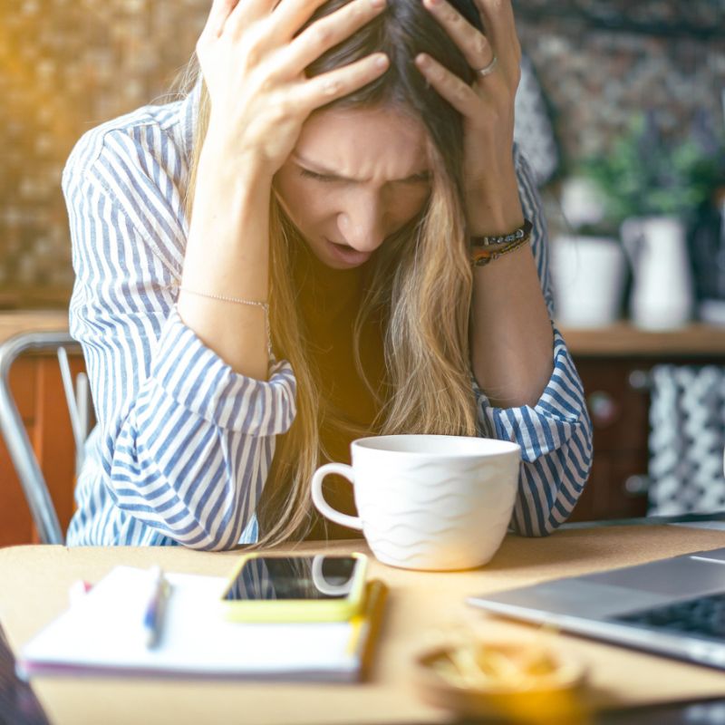 A person in a striped shirt holds their head in their hands, appearing stressed. They sit at a table with a cup, notebook, smartphone, and open laptop, perhaps overwhelmed by researching vaginal yeast infection symptoms.