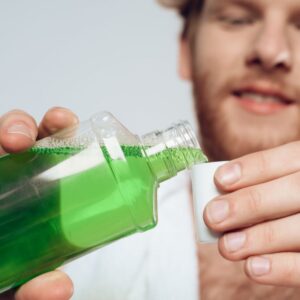A man pours green mouthwash from a clear plastic bottle into a small white cap, mindful to keep his oral health in check and potentially reduce Candida symptoms.