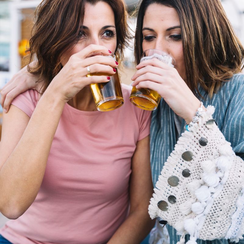 Two women drinking beer from glasses, sitting closely together. One woman is wearing a pink shirt, and the other is wearing a blue patterned sweater, seemingly oblivious to any worries about what causes jock itch.