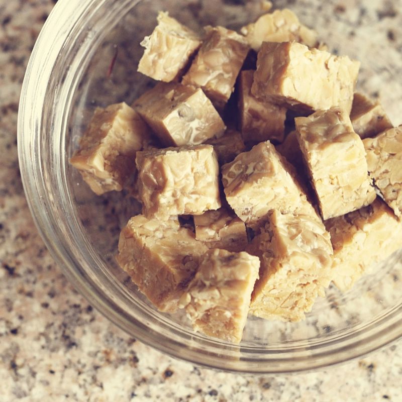 A glass bowl filled with cubed pieces of beige tempeh on a granite countertop.