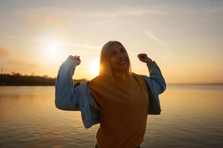 A person stands by a body of water at sunset, smiling with arms raised, embodying self-efficacy while wearing a jacket and sweater.