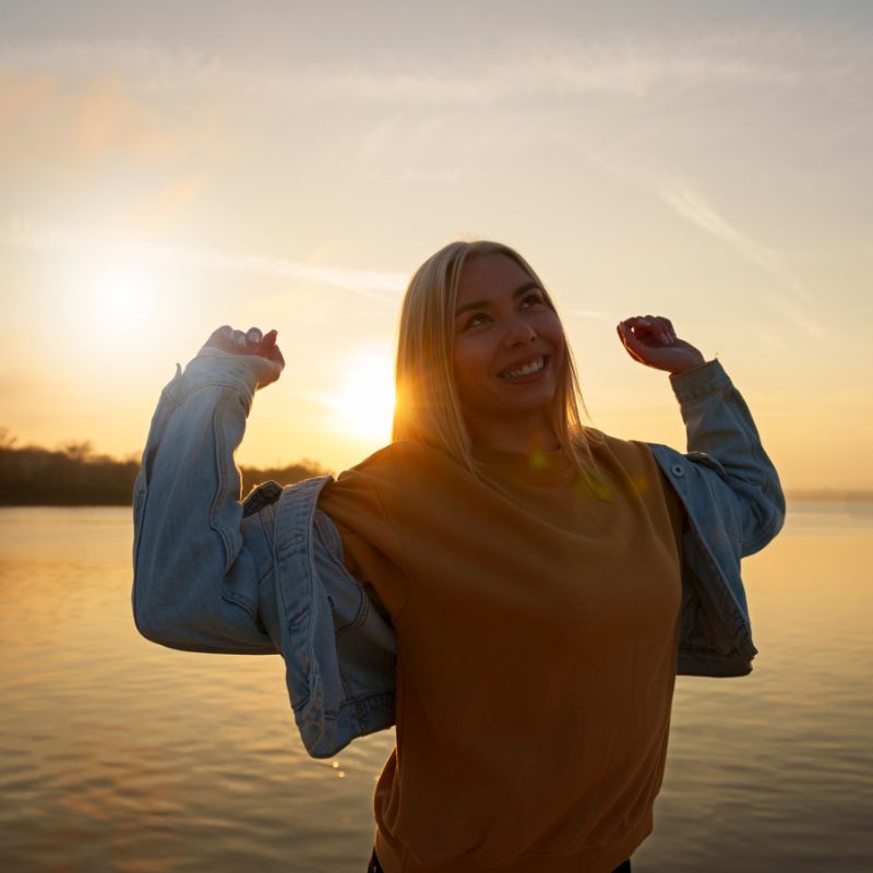 A person with blond hair stands near water at sunset, smiling and lifting their arms in a display of self-efficacy while wearing a yellow shirt and a denim jacket.