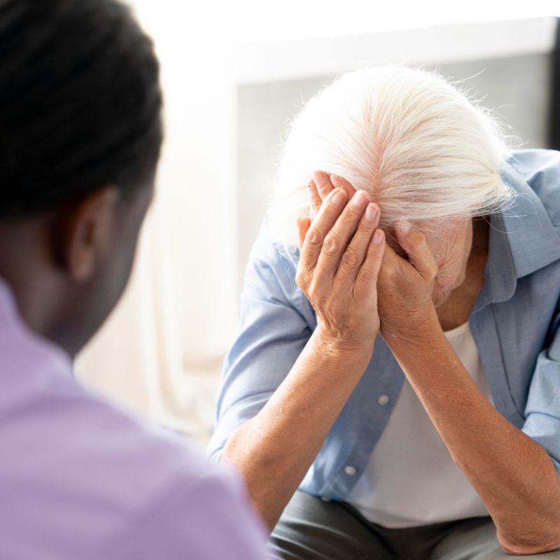 An elderly person with white hair covers their face with their hands while another person, facing away, listens intently. A nearby table holds a steaming mug of tea sprinkled with cinnamon, hinting at the comforting health benefits of cinnamon enjoyed during their conversation.