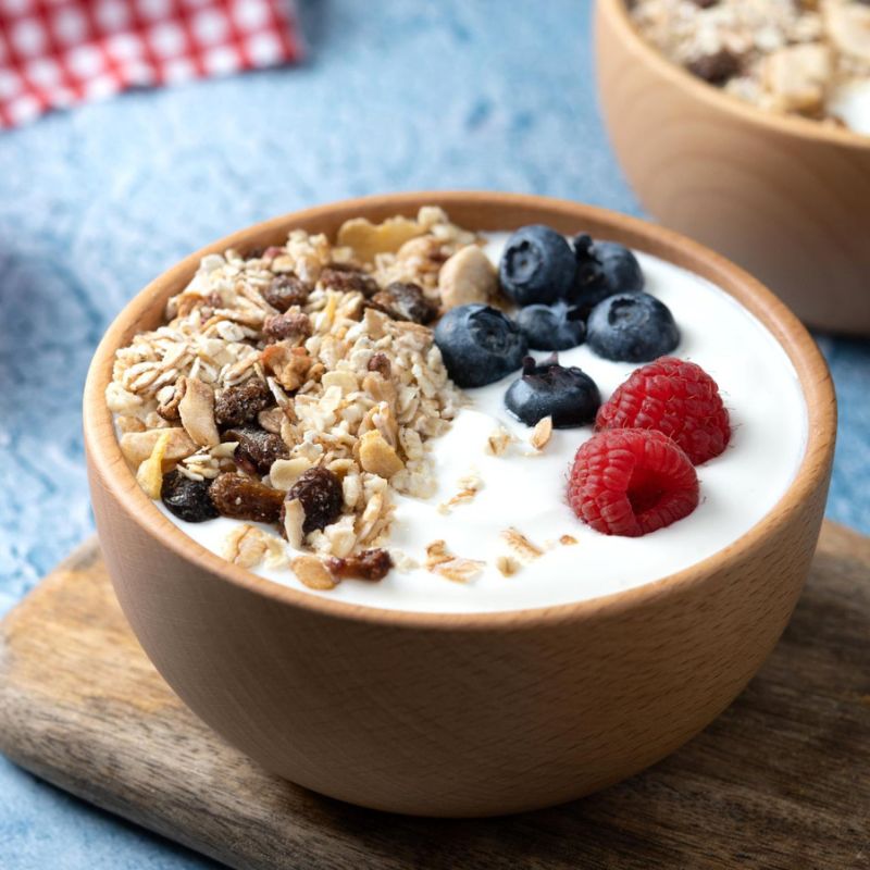 A wooden bowl filled with yogurt, topped with granola, blueberries, and raspberries, placed on a wooden board with a blue surface underneath.