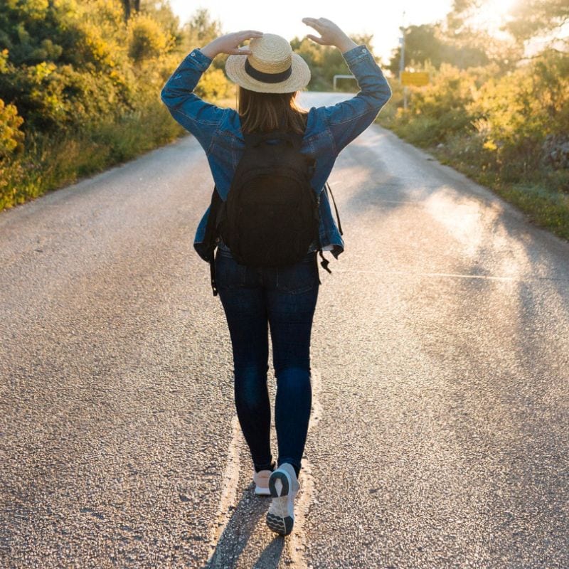 Woman walking down a country road at sunset, holding onto her hat.