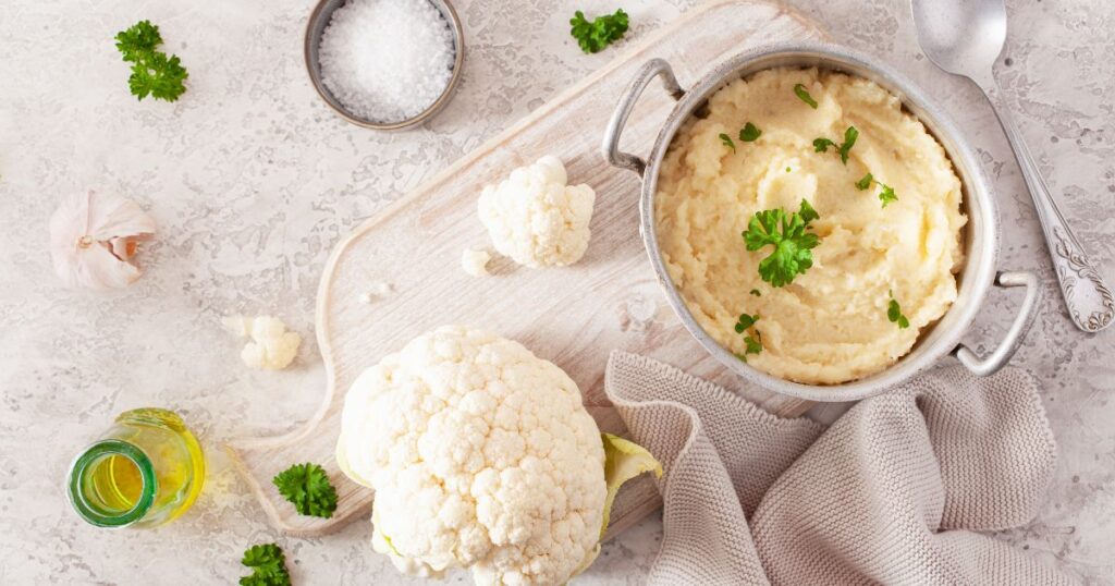 a bowl of herbed cauliflower mash rests on the table with a head of cauliflower and some salt alongside