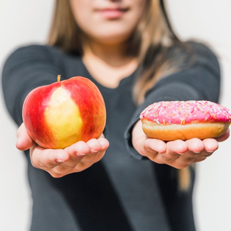 A person holding out a fresh apple in one hand and a frosted donut in the other, symbolizing a choice between healthy and indulgent food options.