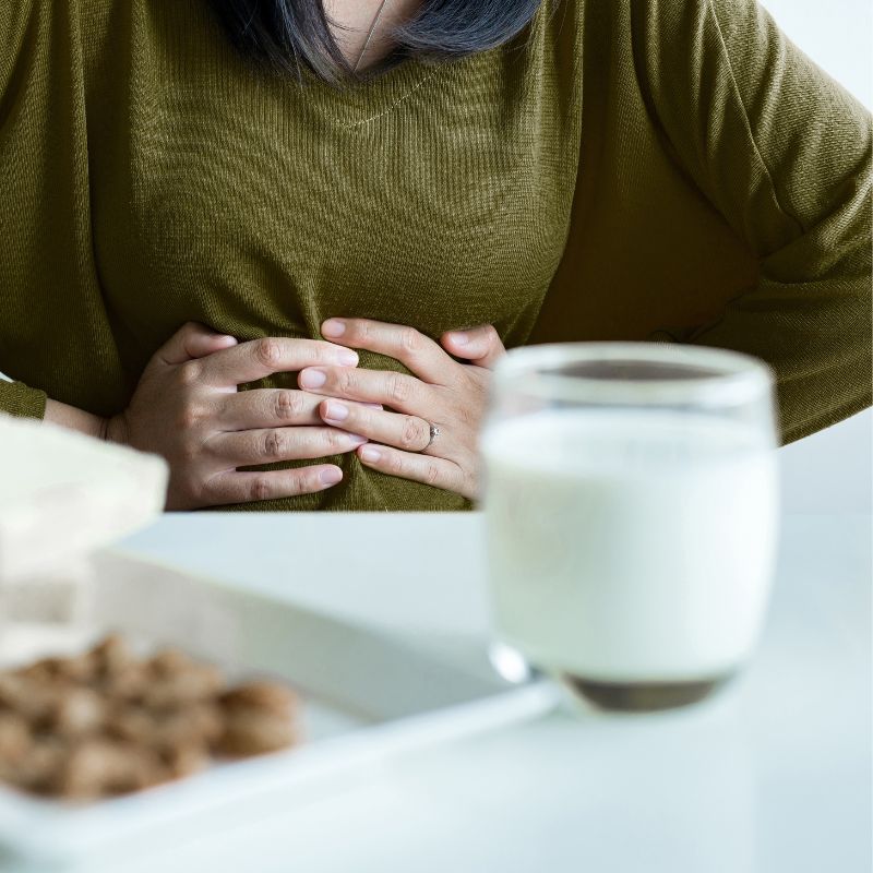 A person clutching their stomach sits at a table with a glass of milk and a plate of cookies in the foreground.