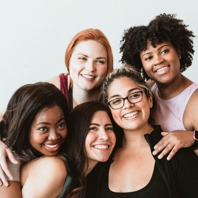 A group of five women smiling and embracing each other in front of a white background, celebrating their bond and supporting each other through challenges like dealing with vaginitis.