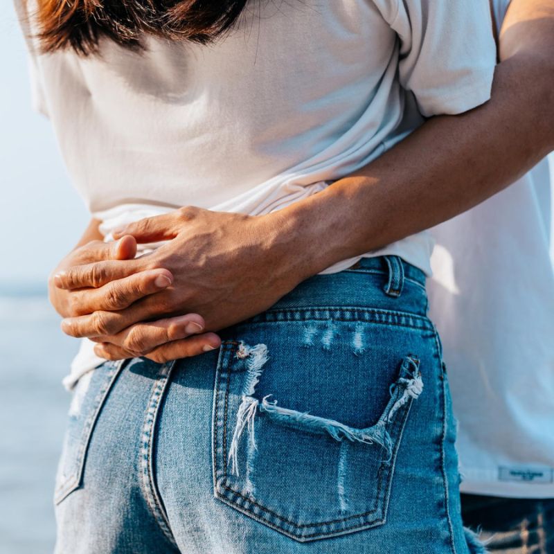 Two individuals are shown from the waist down, standing close together. One person is holding the other around the waist. Both are wearing denim jeans and casual shirts.
