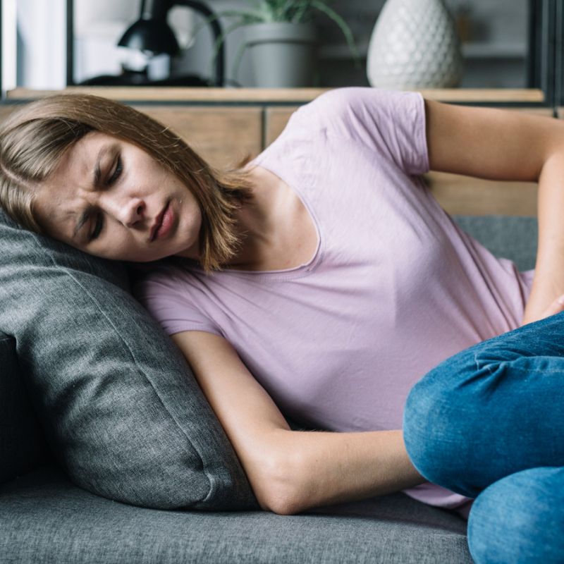 A woman in a light pink shirt lies on a sofa holding her stomach with a pained expression, possibly pondering what causes vaginal yeast infections.