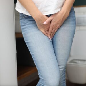 Person in jeans with hands clasped over midsection, standing in front of a toilet, suggesting discomfort or urgency.