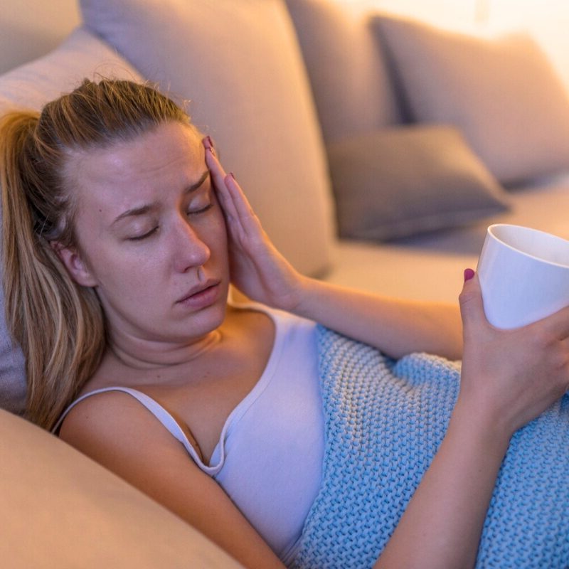 A woman resting on a sofa with a pained expression while holding her head and a cup.