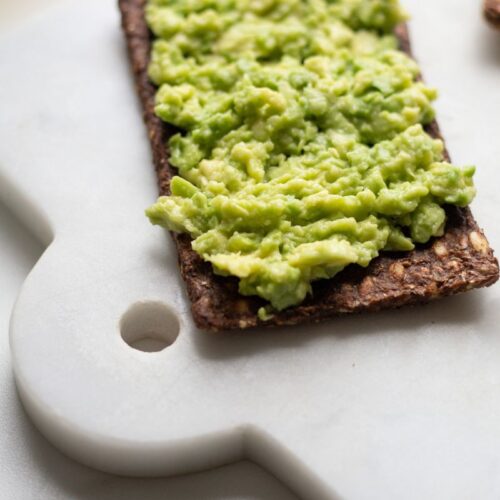 smashed avocado on seed crackers rest on a white marble cutting board