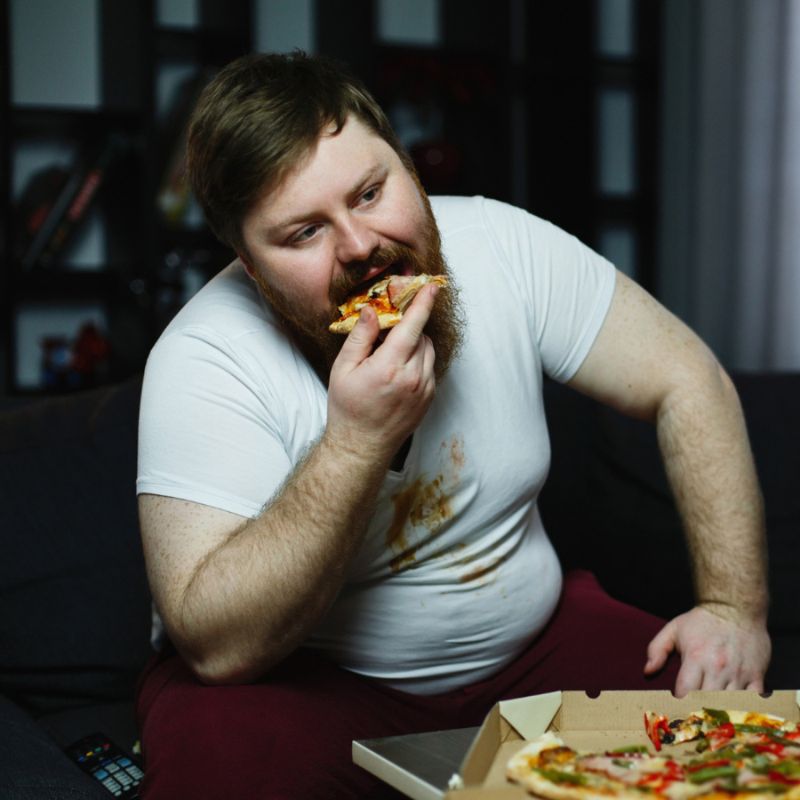 A man in a white shirt with stains is sitting on a couch, eating pizza from a box.