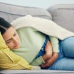 A woman resting on a couch, appearing uncomfortable or in pain, holding her abdomen.