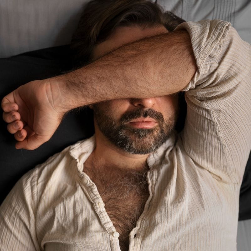 Man with beard covering his eyes with his arm while lying down.